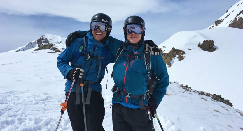 Two people wearing winter gear smile for a photo in front of a mountainous snowy landscape.